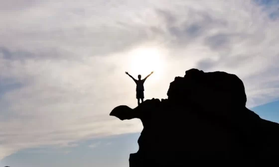 A person triumphantly stands on a mountain peak, arms raised high. They exude joy and accomplishment.