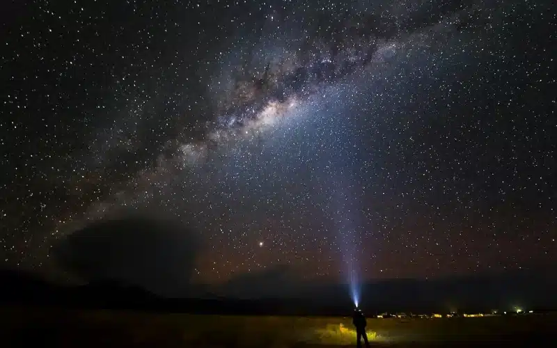 Image of a man stands before a starry sky, holding a flashlight. Trust your heart, embrace its wisdom, and persist, even in darkness